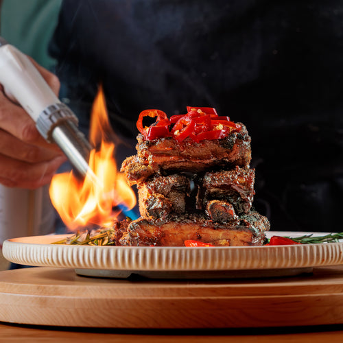 Chef torching stacked barbecue ribs with flames and red peppers on top