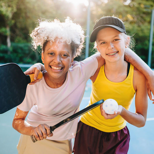 Two young pickleball players smiling together on the courts at The Fort in Fort Lauderdale holding paddles and a pickleball.